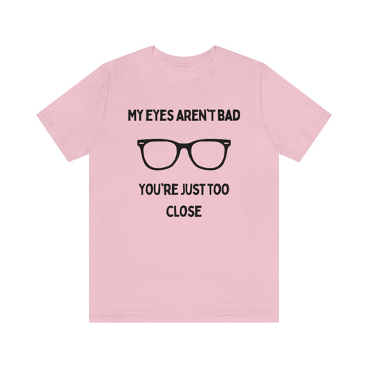 A pink t-shirt with black text reading "My eyes aren't bad, you're just too close" with a pair of glasses in the middle.