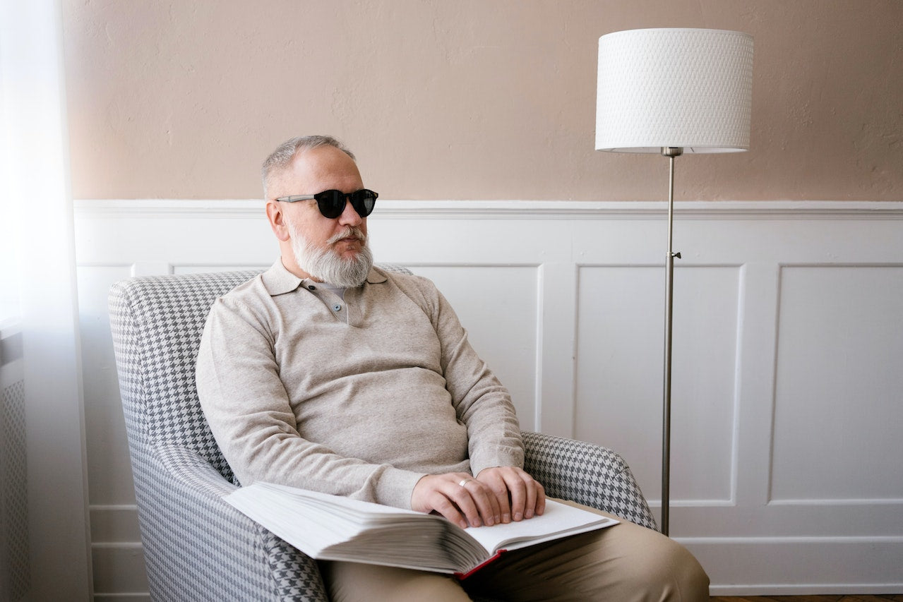 A blind bearded man sitting in a chair reading a book in braille