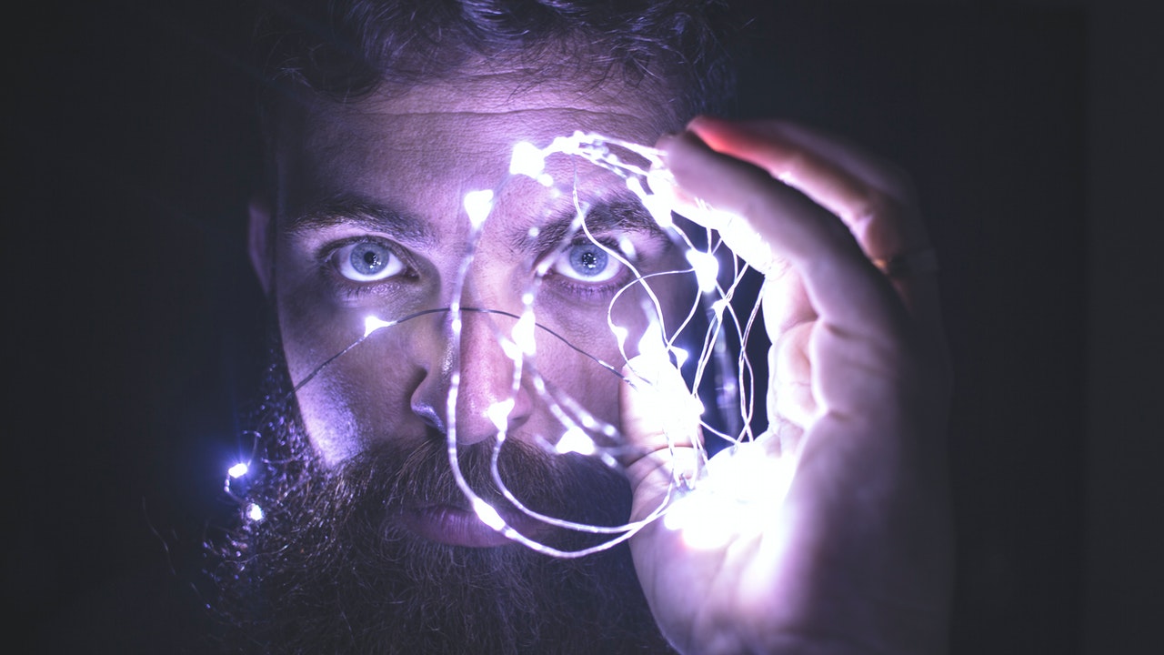 The face of a bearded man holding a string of ironwire with lights on it, glowing purple
