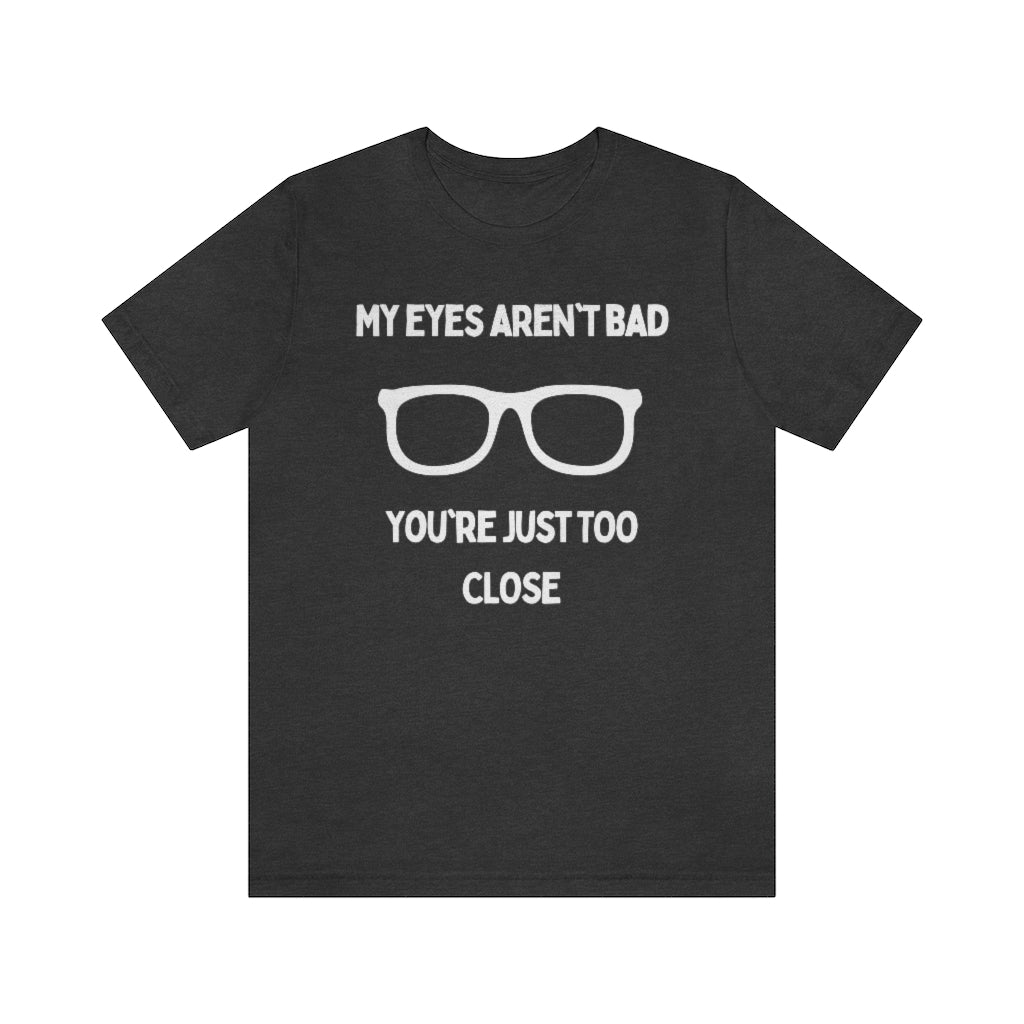 A dark grey heather t-shirt with white text reading "My eyes aren't bad, you're just too close" with a pair of glasses in the middle.