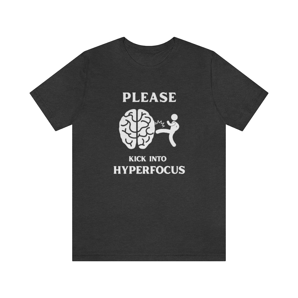 A dark grey heather t-shirt with white text saying "Please kick into hyperfocus". The world 'Please" at the top and inbetween the text is a stickfigure kicking a brain.