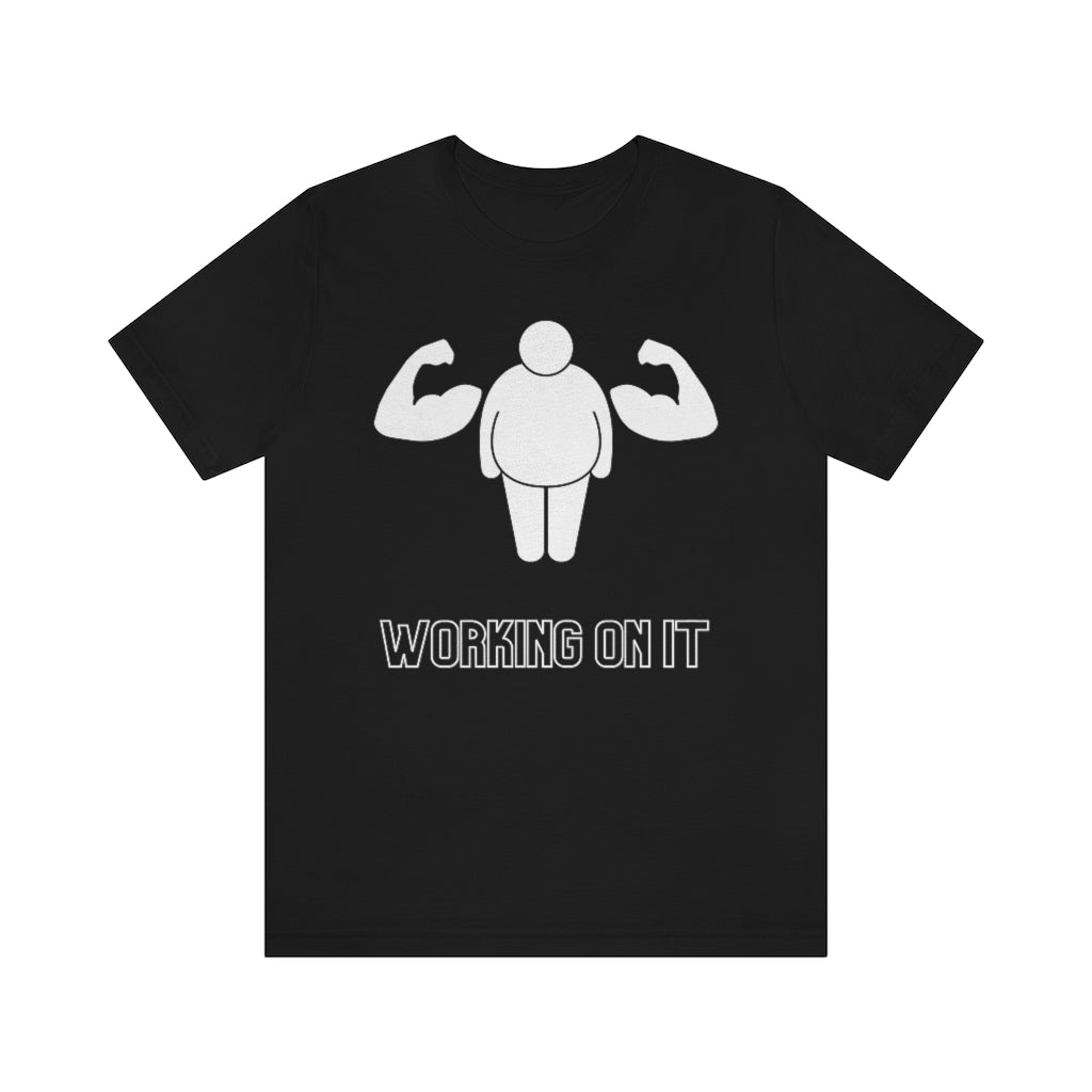 A black t-shirt showing a fat stickfigure with an extra set of  muscular arms. Under it reads: "Working on it".