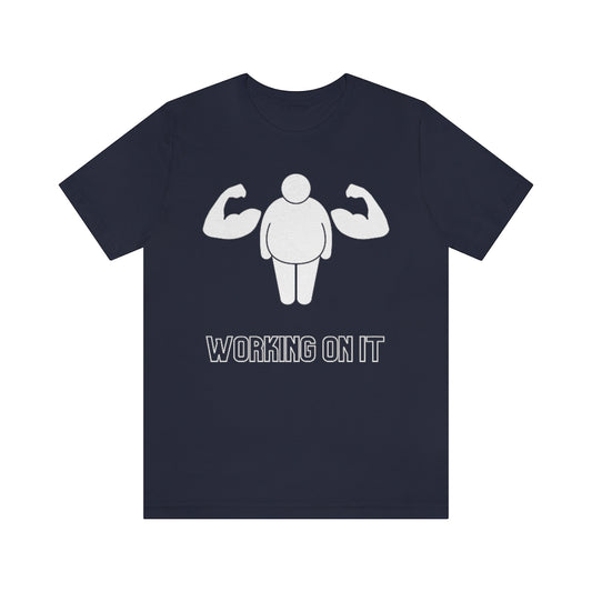 A navy colored t-shirt showing a fat stickfigure with an extra set of muscular arms. Under it reads: "Working on it".