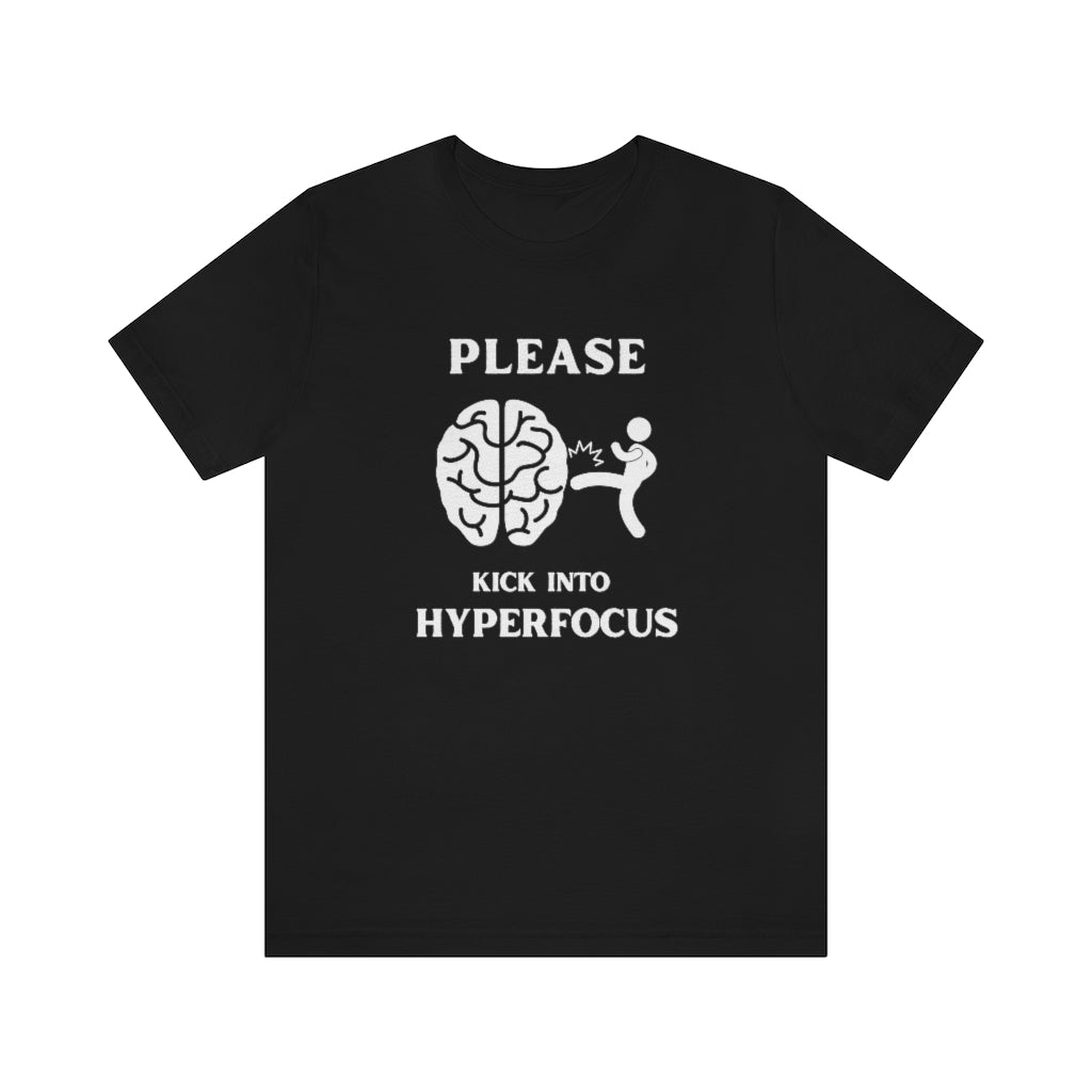 A black t-shirt with white text saying "Please kick into hyperfocus". The world 'Please" at the top and inbetween the text is a stickfigure kicking a brain.