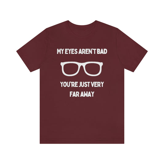 A maroon-colored t-shirt with white text reading "My eyes aren't bad, you're just very far away" with a pair of glasses in the middle.