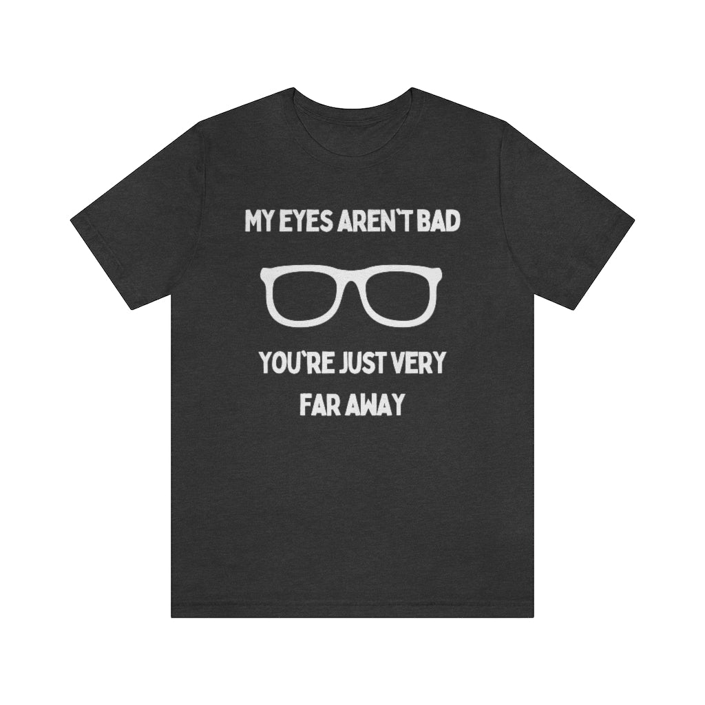 A dark grey heather t-shirt with white text reading "My eyes aren't bad, you're just very far away" with a pair of glasses in the middle.