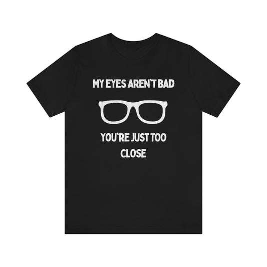 A black t-shirt with white text reading "My eyes aren't bad, you're just too close" with a pair of glasses in the middle.