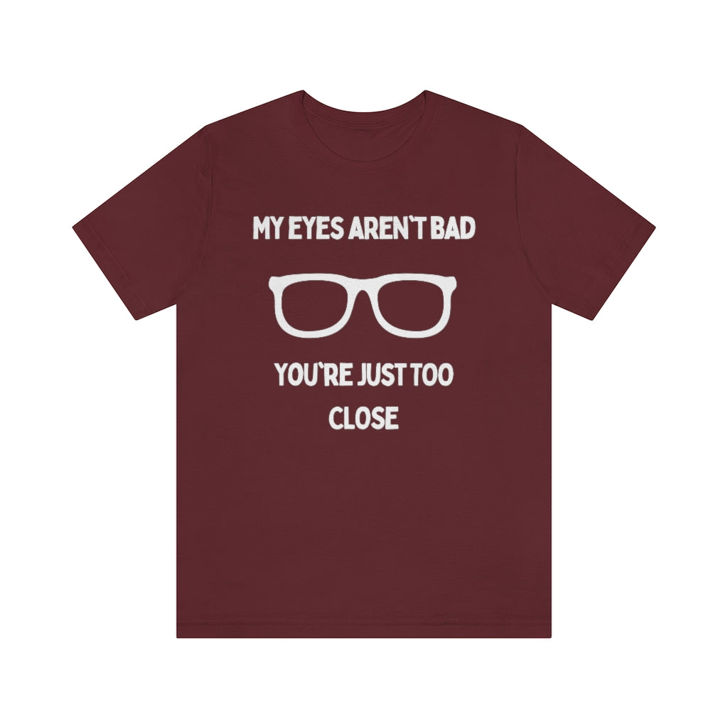 A maroon-colored t-shirt with white text reading "My eyes aren't bad, you're just too close" with a pair of glasses in the middle.