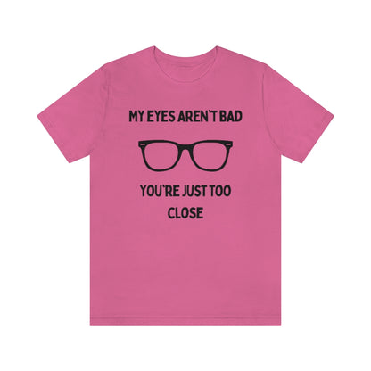 A charity pink t-shirt with black text reading "My eyes aren't bad, you're just too close" with a pair of glasses in the middle.