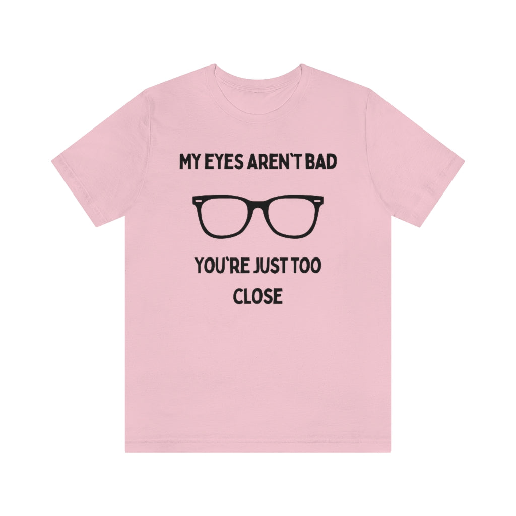 A pink t-shirt with black text reading "My eyes aren't bad, you're just too close" with a pair of glasses in the middle.