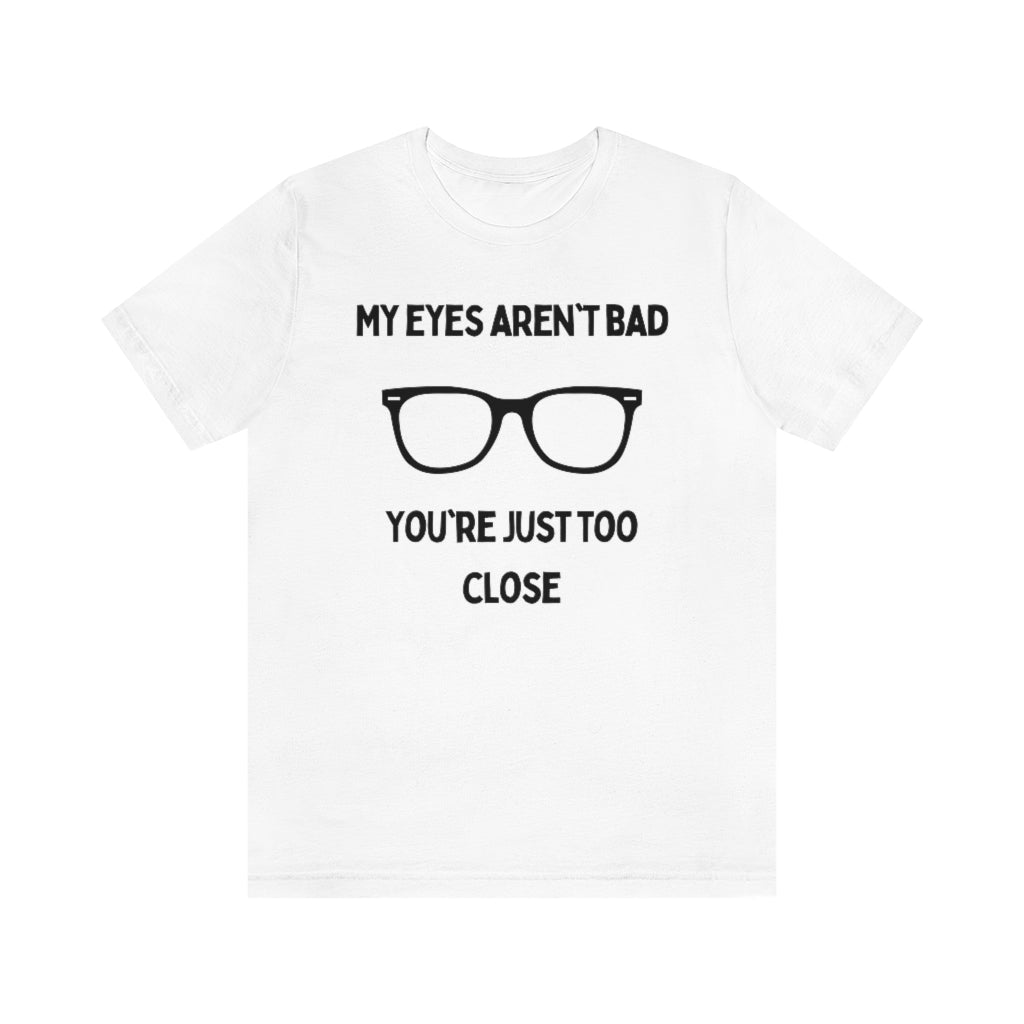 A white t-shirt with black text reading "My eyes aren't bad, you're just too close" with a pair of glasses in the middle.